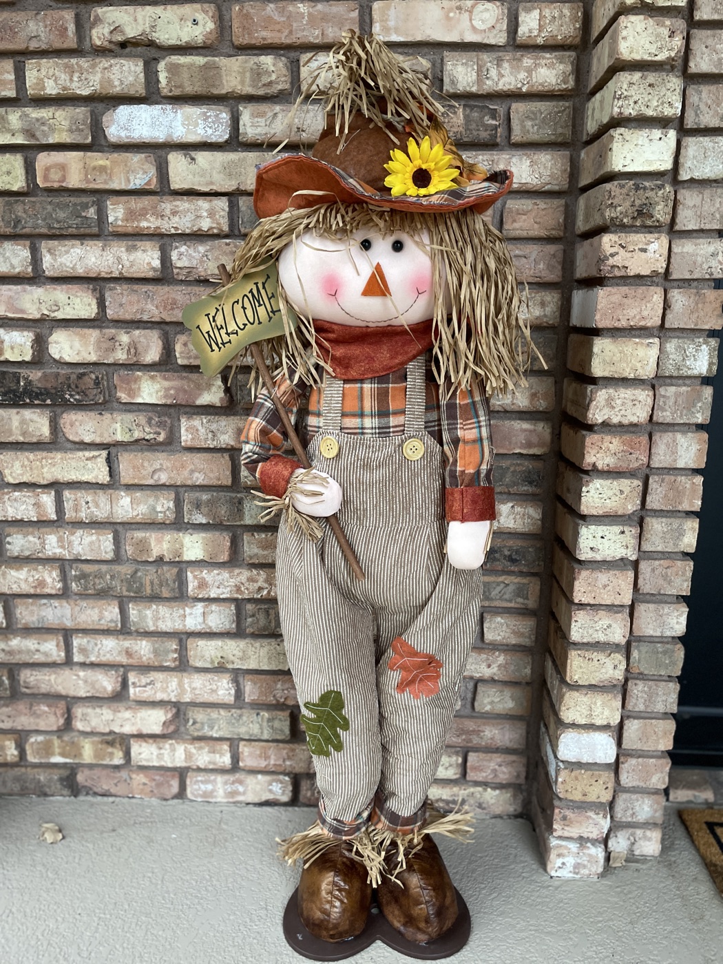 Smiling Welcome Scarecrow in striped overalls and an orange plaid shirt, holding a Welcome sign